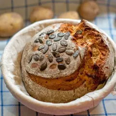 Ein frisch gebackenes Kartoffelbrot mit Kürbiskernen, bereit für das Kartoffelbrot Rezept. Kartoffeln liegen unscharf im Hintergrund.