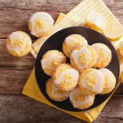 Zitronen-Muffins, frisch aus dem Thermomix, mit Zuckerguss und Zitronenschale bestreut, auf einem Teller mit Zitronenspalten.