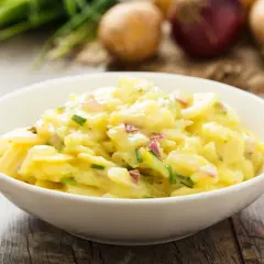 Ein Schüsselchen mit Omas Kartoffelsalat, cremig und mit frischen Kräutern verfeinert. Dazu liegen Kartoffeln und Zwiebeln bereit.