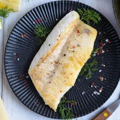Perfekt gebratenes Rotbarschfilet auf schwarzem Teller, garniert mit Dill und bunten Pfefferkörnern. Dazu Zitronenspalten.