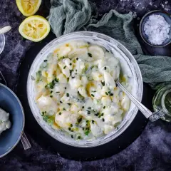 Cremiger Kartoffelsalat mit Joghurt, garniert mit Schnittlauch. Dazu Zitronenspalten und eine kleine Schüssel mit Kräutern.