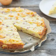 Ein goldbrauner Zwiebelkuchen ohne Boden, mit Speckstücken und einer cremigen Füllung, liegt auf einem Holztisch. Daneben eine Scheibe auf einem Teller und eine Zwiebel.