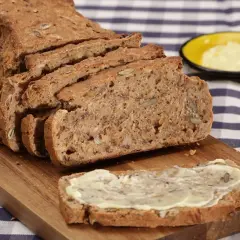 Ein frisch gebackenes Vollkornbrot mit vielen Kernen, aufgeschnitten auf einem Holzbrett. Ein Stück ist mit Butter bestrichen. Perfekt für ein gesundes Frühstück oder Abendbrot.
