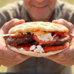 Ein Mann hält eine saftige Pljeskavica mit Tomaten, Zwiebeln und Käse. Ein tolles Rezept für den Sommer!