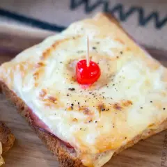Ein klassisches Toast Hawaii mit geschmolzenem Käse, Schinken und einer Kirsche, serviert auf einem Holzbrett.