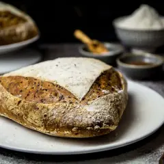Ein rustikales Dinkelbrot mit Leinsamen liegt auf einem Teller, bestäubt mit Mehl. Im Hintergrund eine Schüssel mit Mehl und kleine Schälchen.