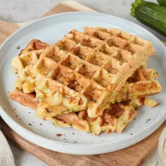 Goldbraune Zucchini-Waffeln auf einem Teller, daneben frische Zucchini und geriebener Käse. Ein leckeres zucchini waffeln rezept.