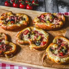 Fünf kleine Brezel Pizza mit Schinken, Oliven und Tomaten, auf Backpapier auf einem Holzbrett angerichtet.