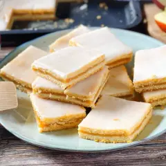 Rechteckige Stückchen Apfelkuchen mit einer zarten Apfelmusfüllung und weißer Glasur, auf einem Teller angerichtet.