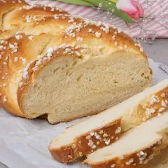 Ein goldbrauner Hefezopf, bestreut mit Zuckerkristallen, liegt auf Backpapier. Ein Stück ist angeschnitten, daneben liegen drei Scheiben. Ein tolles Rezept für Hefezopf!