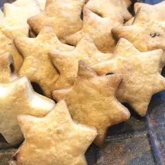 Ein Haufen goldbrauner Dinkelkekse ohne Zucker in Sternform, daneben Zimtstangen auf einem rustikalen Teller.