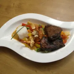 Zwei saftige Rinderfiletstücke auf einem Bett aus Gemüse und Garnelen, serviert mit Reis in einer cremigen Sauce, die an einen **thai curry** erinnert.