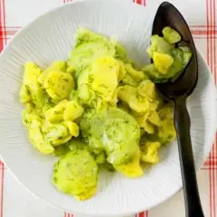 Ein Teller mit Kartoffelsalat mit Gurken und Dill, garniert mit frischem Dill. Ein Löffel mit dem Salat liegt auf dem Teller.