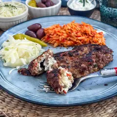 Gefülltes Bifteki-Rezept auf einem Teller mit Reisnudeln, Krautsalat, Oliven und Peperoni. Ein Stück wird mit einer Gabel angeschnitten.