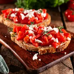 Zwei rustikale Scheiben Brot belegt mit frischen Tomatenwürfeln und zerbröseltem Feta. Bruschetta mit Feta, garniert mit Kräutern, auf einem Holzbrett.