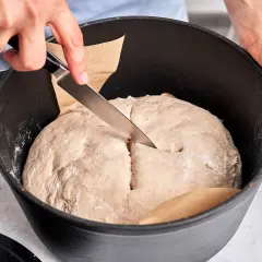 Hände schneiden mit Messer in den Teig für ein "friss dich dumm Brot" in einem Gusseisentopf.