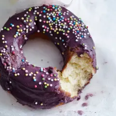 Ein lila glasierter Donut mit bunten Streuseln, ein Bissen fehlt. Perfekt für ein schnelles Donuts Rezept.
