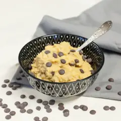 Schüssel mit essbarem Keksteig und Schokostückchen, daneben verstreute Schokotropfen. Ein Löffel steckt im Teig.