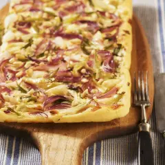 Flammkuchen mit roten Zwiebeln, Pinienkernen und Käse auf einem Holzbrett, daneben Gabel und Messer.
