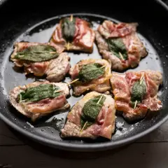 Saltimbocca alla romana in einer Pfanne, umgeben von Kräutern und Gewürzen.