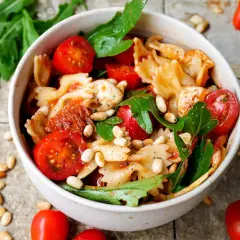 Schüssel mit Nudelsalat, Farfalle, Kirschtomaten, Mozzarella, Pinienkernen und Rucola. Ein köstlicher nudelsalat mit getrockneten tomaten.