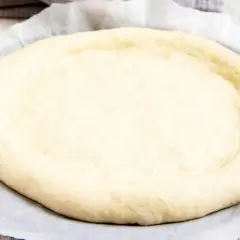 Ein runder, heller pizzateig mit trockenhefe liegt auf Backpapier. Die Ränder sind leicht angehoben.