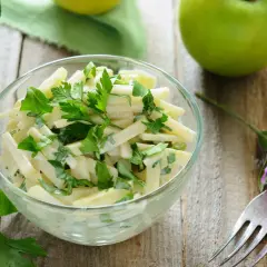 Frischer Kohlrabi Salat mit Petersilie in einer Glasschüssel, garniert mit einer lila Blüte und grünen Äpfeln im Hintergrund.