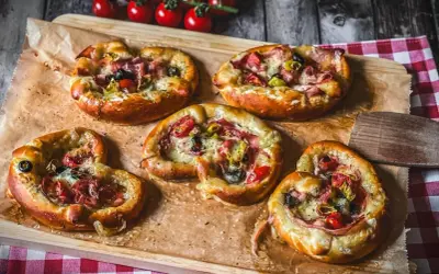 Fünf kleine Brezel Pizza mit Schinken, Oliven und Tomaten, auf Backpapier auf einem Holzbrett angerichtet.