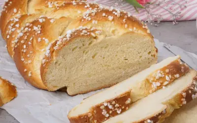 Ein goldbrauner Hefezopf, bestreut mit Zuckerkristallen, liegt auf Backpapier. Ein Stück ist angeschnitten, daneben liegen drei Scheiben. Ein tolles Rezept für Hefezopf!