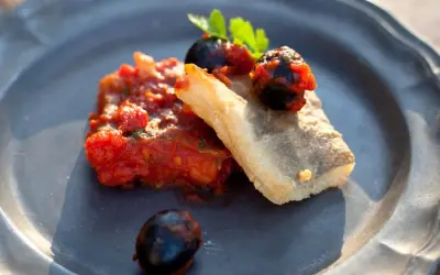 Baccalà alla livornese: ein Stück gebratenes Kabeljaufilet mit Tomaten, Oliven und Petersilie auf einem grauen Teller.