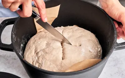 Hände schneiden mit Messer in den Teig für ein "friss dich dumm Brot" in einem Gusseisentopf.