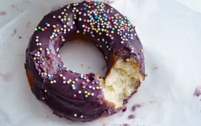 Ein lila glasierter Donut mit bunten Streuseln, ein Bissen fehlt. Perfekt für ein schnelles Donuts Rezept.