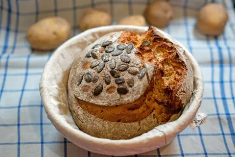 Ein frisch gebackenes Kartoffelbrot mit Kürbiskernen, bereit für das Kartoffelbrot Rezept. Kartoffeln liegen unscharf im Hintergrund.
