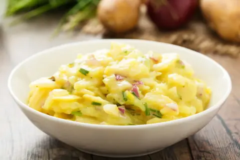 Ein Schüsselchen mit Omas Kartoffelsalat, cremig und mit frischen Kräutern verfeinert. Dazu liegen Kartoffeln und Zwiebeln bereit.