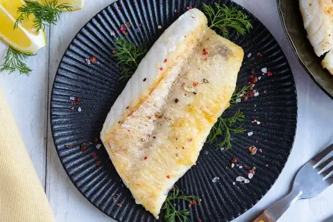 Perfekt gebratenes Rotbarschfilet auf schwarzem Teller, garniert mit Dill und bunten Pfefferkörnern. Dazu Zitronenspalten.