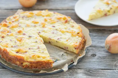 Ein goldbrauner Zwiebelkuchen ohne Boden, mit Speckstücken und einer cremigen Füllung, liegt auf einem Holztisch. Daneben eine Scheibe auf einem Teller und eine Zwiebel.