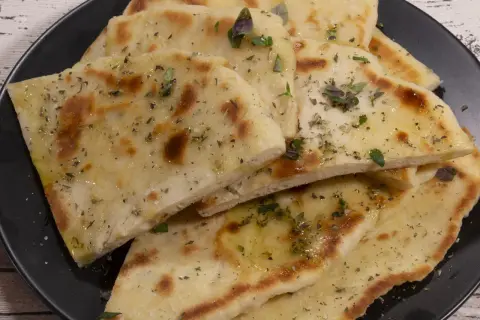 Griechisches Fladenbrot mit Trockenhefe, bestreut mit Kräutern und Olivenöl, auf einem schwarzen Teller.