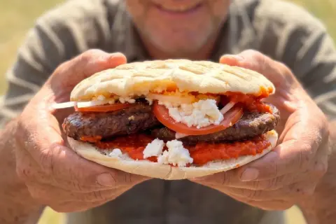 Ein Mann hält eine saftige Pljeskavica mit Tomaten, Zwiebeln und Käse. Ein tolles Rezept für den Sommer!
