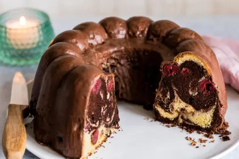 Ein saftiger Marmorkuchen mit Kirschen, überzogen mit Schokoladenglasur, auf einem weißen Teller.