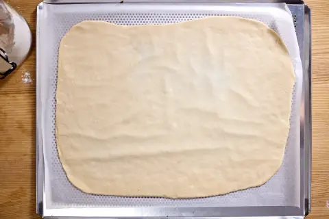 Dünn ausgerollter Flammkuchenteig liegt auf einem Backblech, bereit für leckere Beläge.