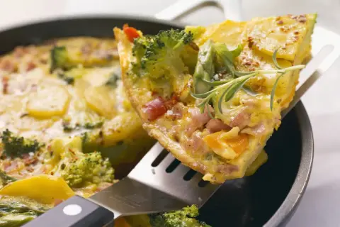 Ein Stück herzhaftes Omelett mit Brokkoli, Kartoffeln und Schinken wird aus einer Pfanne gehoben. Ein tolles Omelett Rezept für jeden Tag.