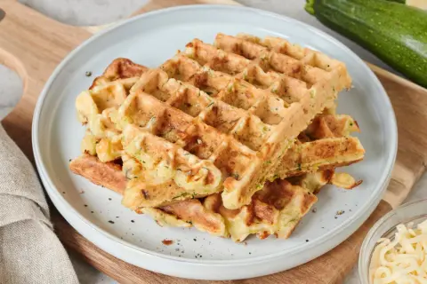 Goldbraune Zucchini-Waffeln auf einem Teller, daneben frische Zucchini und geriebener Käse. Ein leckeres zucchini waffeln rezept.