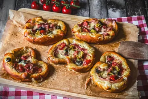 Fünf kleine Brezel Pizza mit Schinken, Oliven und Tomaten, auf Backpapier auf einem Holzbrett angerichtet.