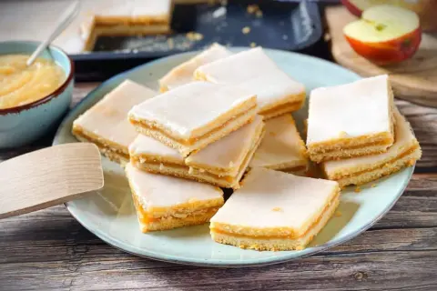 Rechteckige Stückchen Apfelkuchen mit einer zarten Apfelmusfüllung und weißer Glasur, auf einem Teller angerichtet.