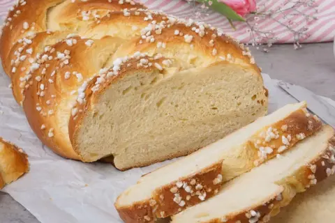 Ein goldbrauner Hefezopf, bestreut mit Zuckerkristallen, liegt auf Backpapier. Ein Stück ist angeschnitten, daneben liegen drei Scheiben. Ein tolles Rezept für Hefezopf!