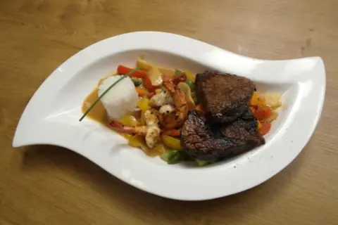 Zwei saftige Rinderfiletstücke auf einem Bett aus Gemüse und Garnelen, serviert mit Reis in einer cremigen Sauce, die an einen **thai curry** erinnert.