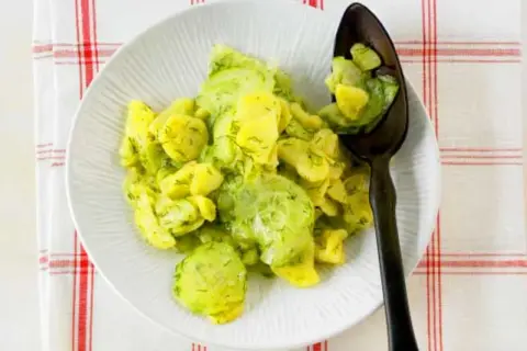 Ein Teller mit Kartoffelsalat mit Gurken und Dill, garniert mit frischem Dill. Ein Löffel mit dem Salat liegt auf dem Teller.