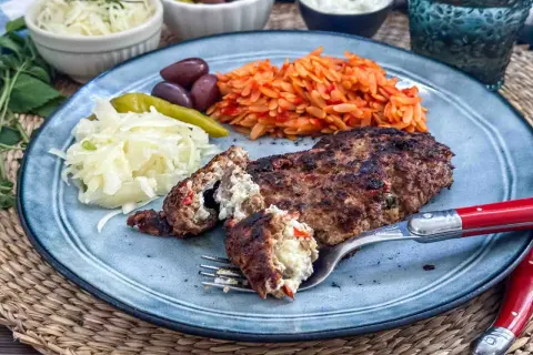 Gefülltes Bifteki-Rezept auf einem Teller mit Reisnudeln, Krautsalat, Oliven und Peperoni. Ein Stück wird mit einer Gabel angeschnitten.