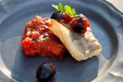 Baccalà alla livornese: ein Stück gebratenes Kabeljaufilet mit Tomaten, Oliven und Petersilie auf einem grauen Teller.