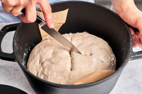 Hände schneiden mit Messer in den Teig für ein "friss dich dumm Brot" in einem Gusseisentopf.